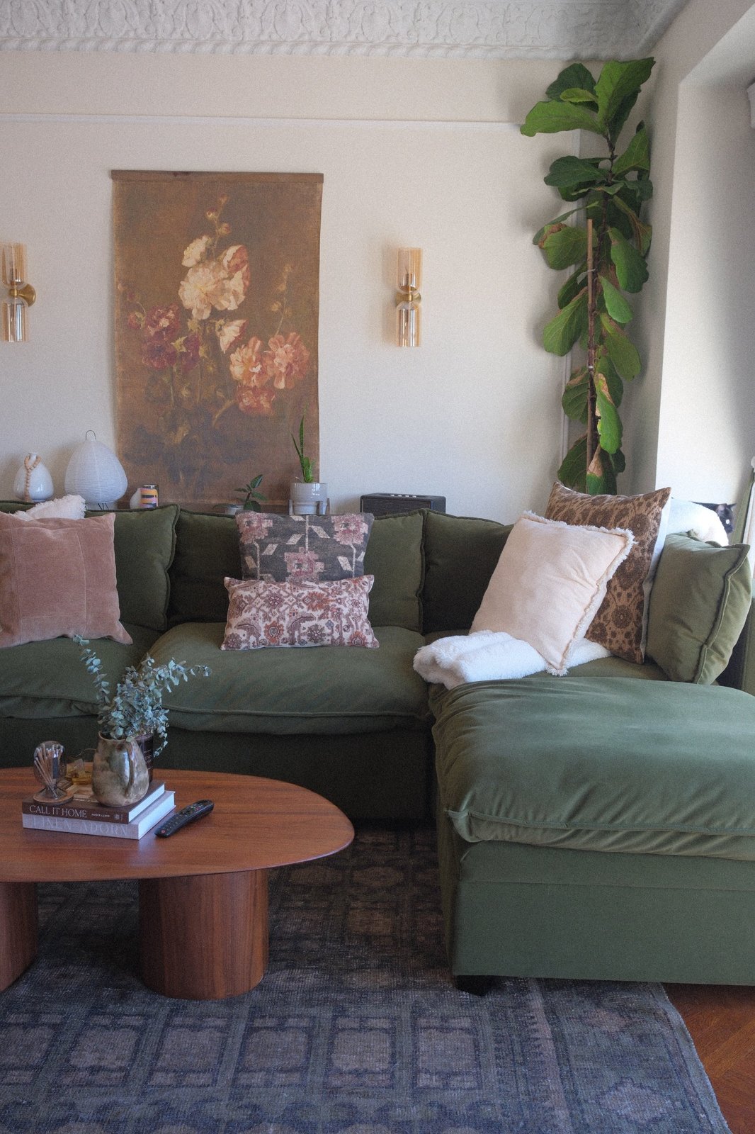 Olive velvet sectional under a vintage floral wall hanging, with amber sconces and a tall fiddle-leaf fig