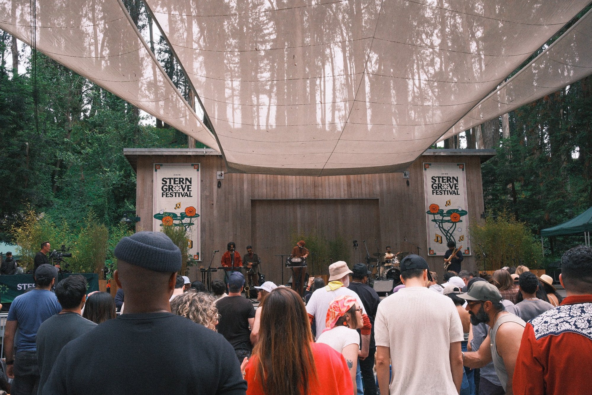 Stern Grove Festival: the crowd under a sail canopy patterned with tree shadows, band on the wooden stage