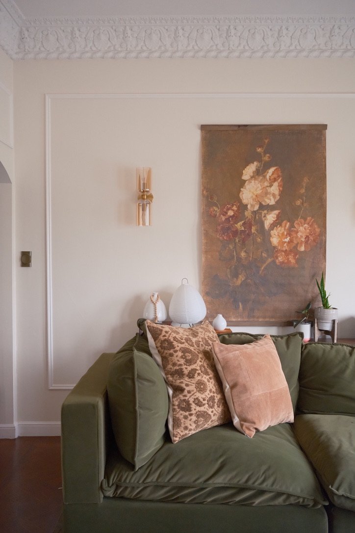 Olive velvet sofa with patterned pillows beneath a vintage floral wall hanging