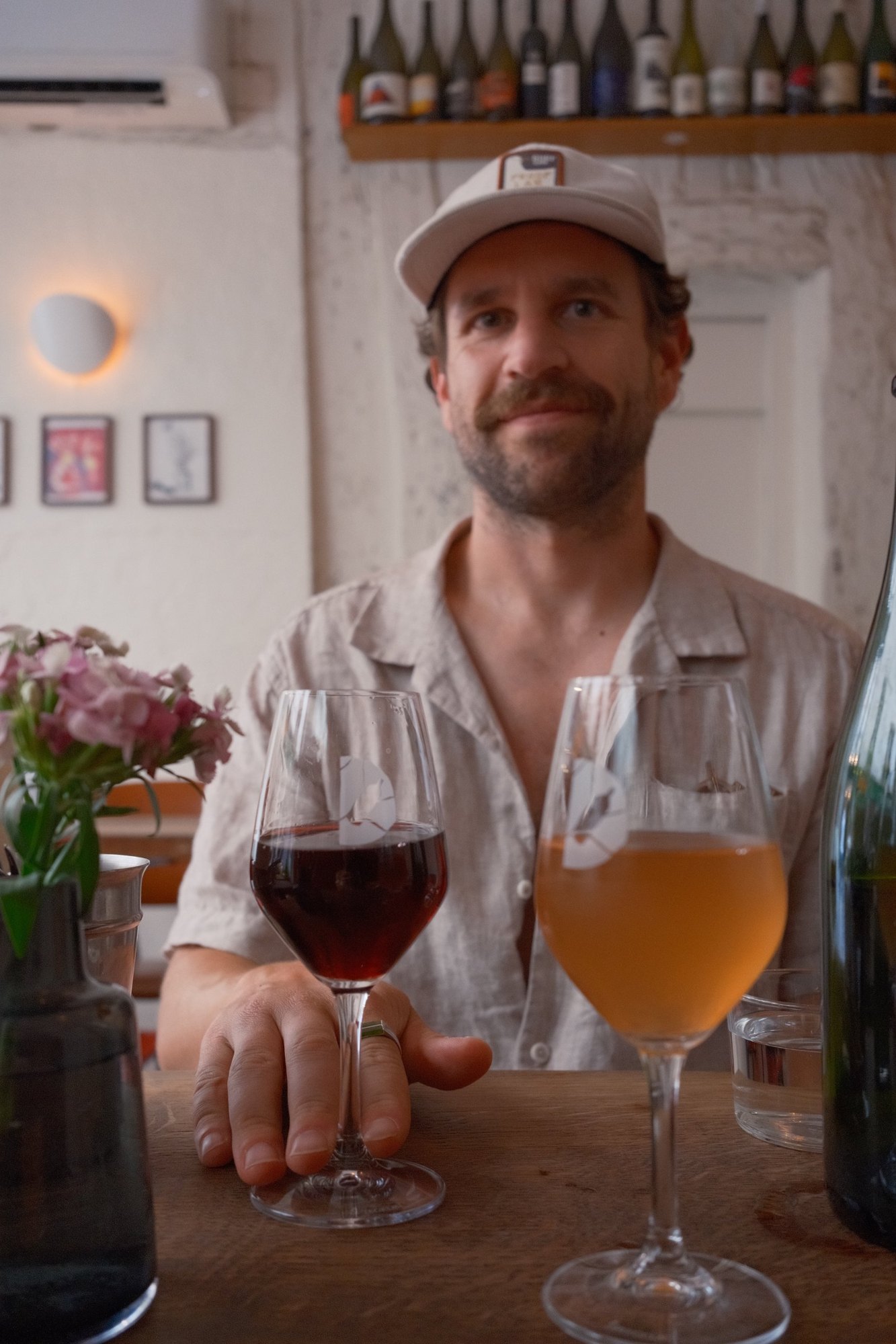 Red and orange natural wine across a wooden table, a smile behind the glasses