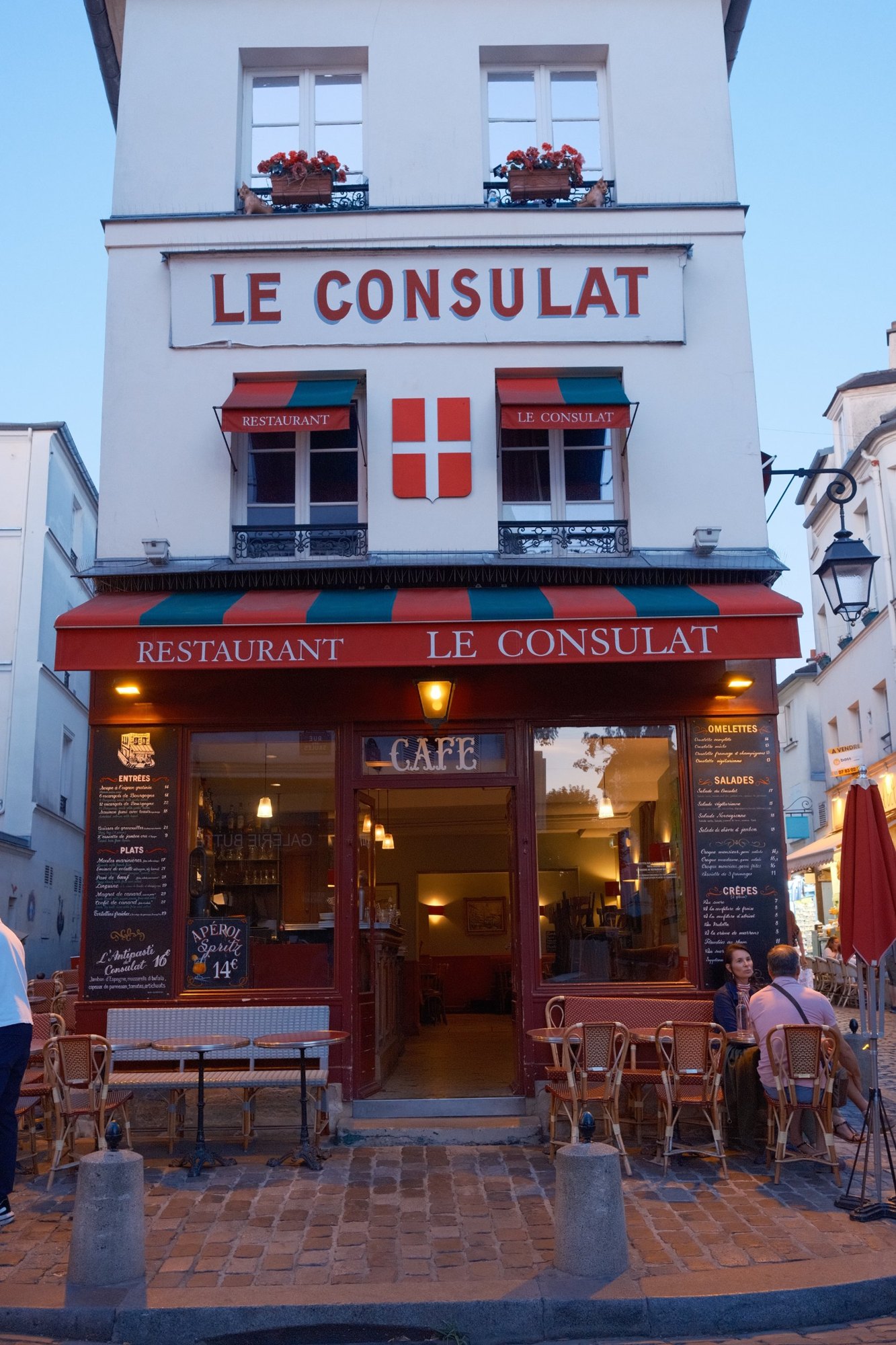 Le Consulat in blue hour: red-and-green awnings, cane chairs and chalkboard menus