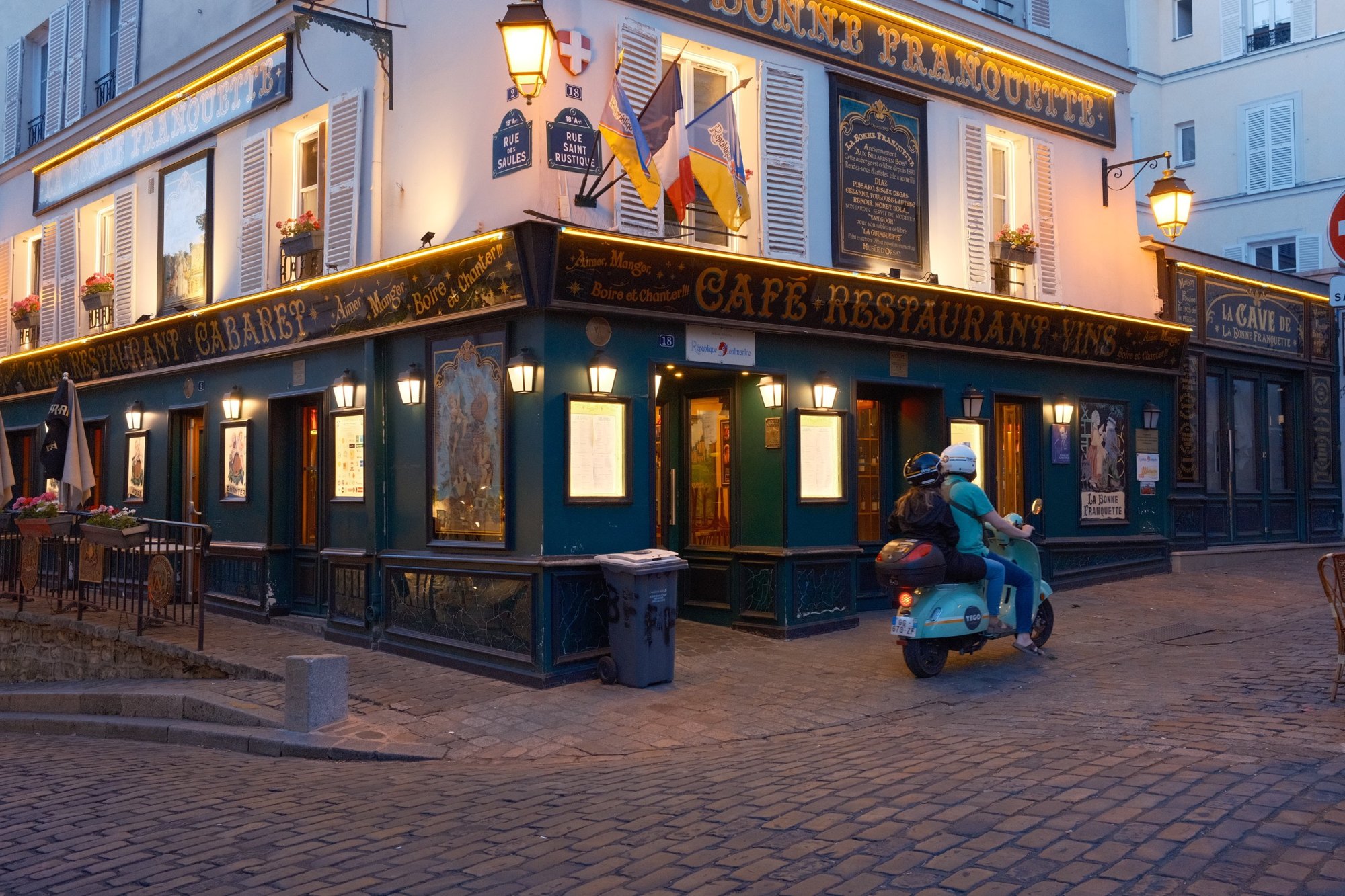 A couple on a mint scooter rounding the glowing corner of La Bonne Franquette at dusk