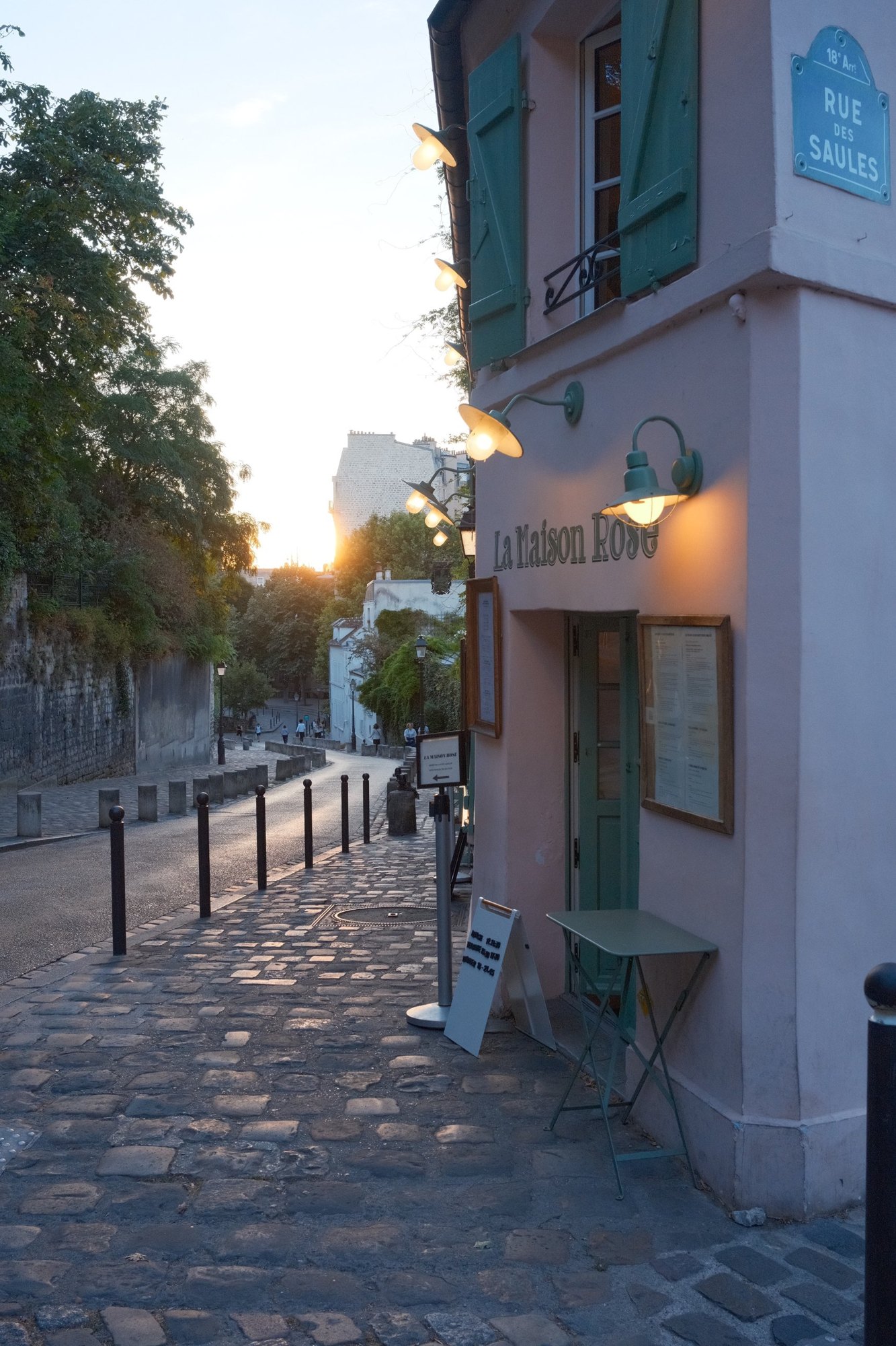 La Maison Rose's lamps glowing at dusk, the street falling away into the sun