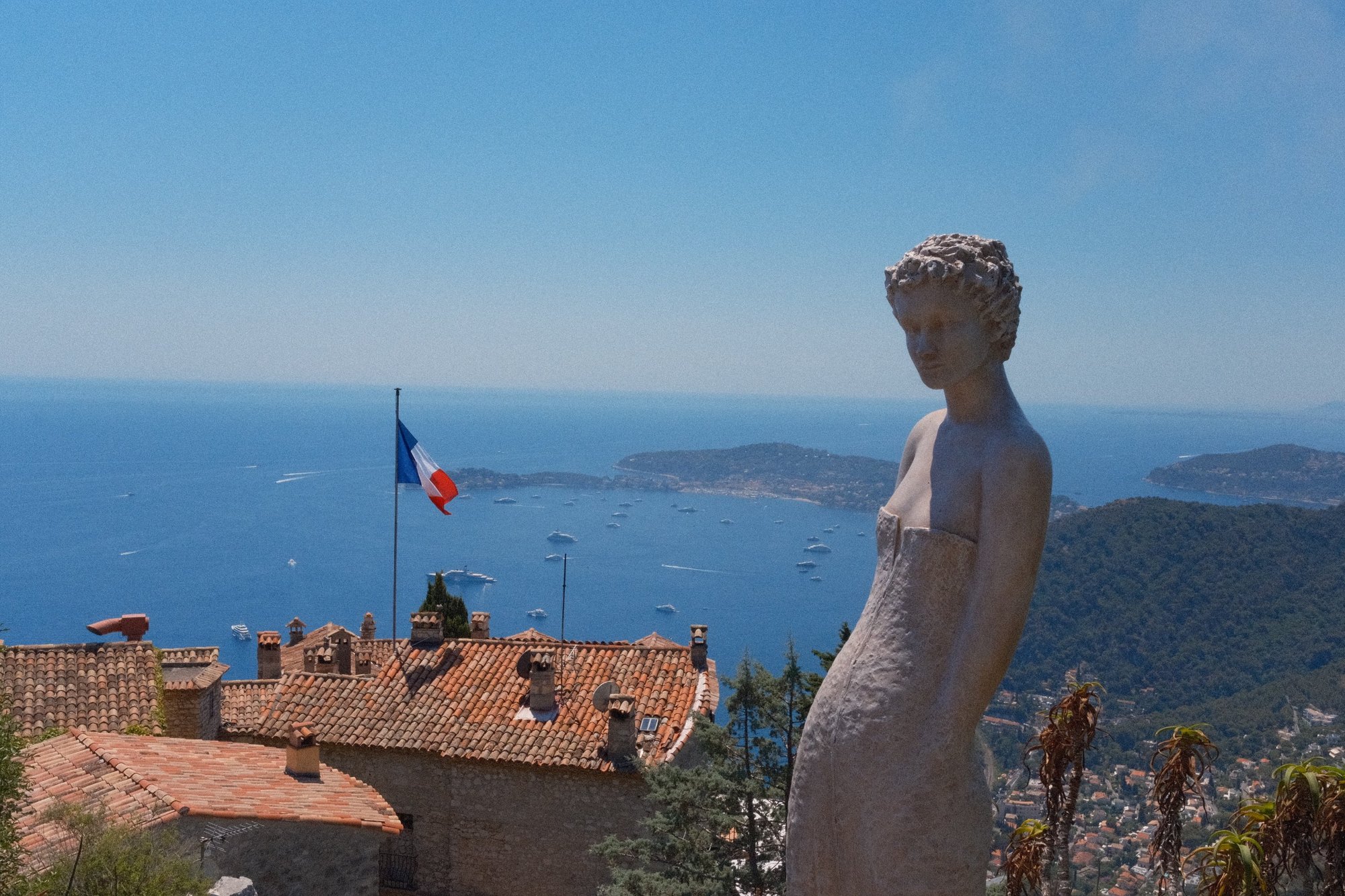A white statue above terracotta roofs and the yacht-filled bay below Èze