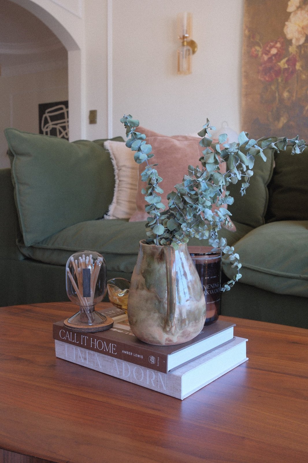 Close-up of a glazed ceramic pitcher with eucalyptus on books, the velvet sofa softly behind