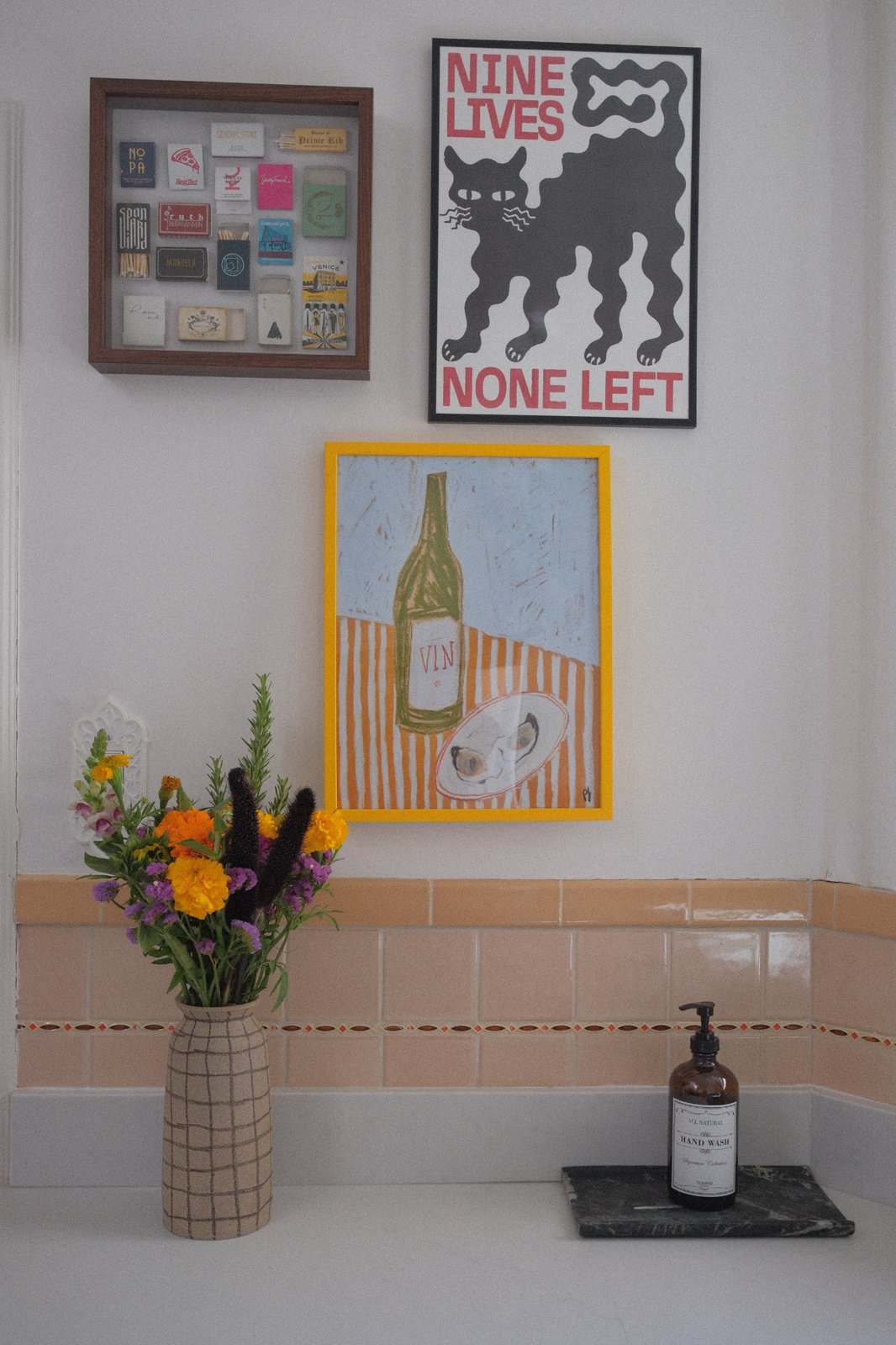 Gallery wall of a matchbook shadow box and graphic prints above peach vintage tile and a grid-painted vase