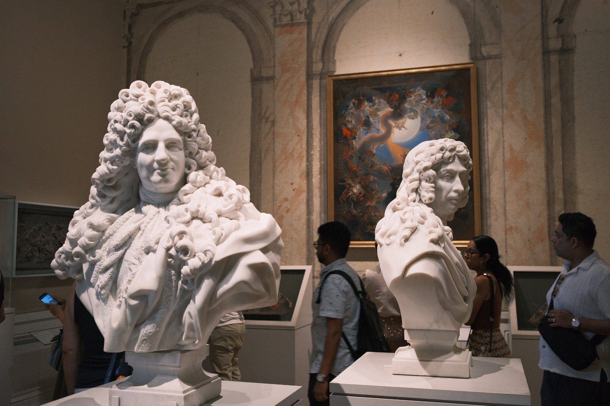 Two marble busts in a museum gallery, a swirling painted ceiling study behind