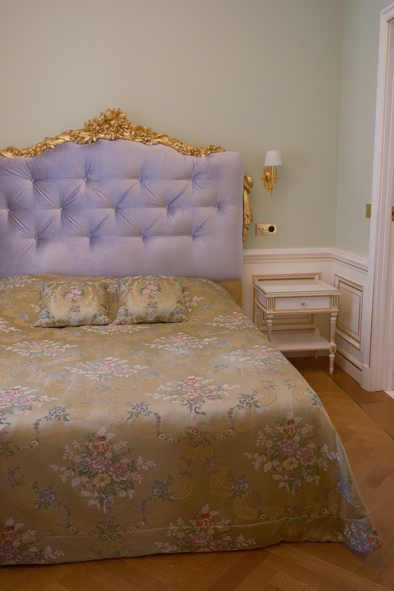 A lavender tufted headboard with a gilded crest over a floral brocade bedspread