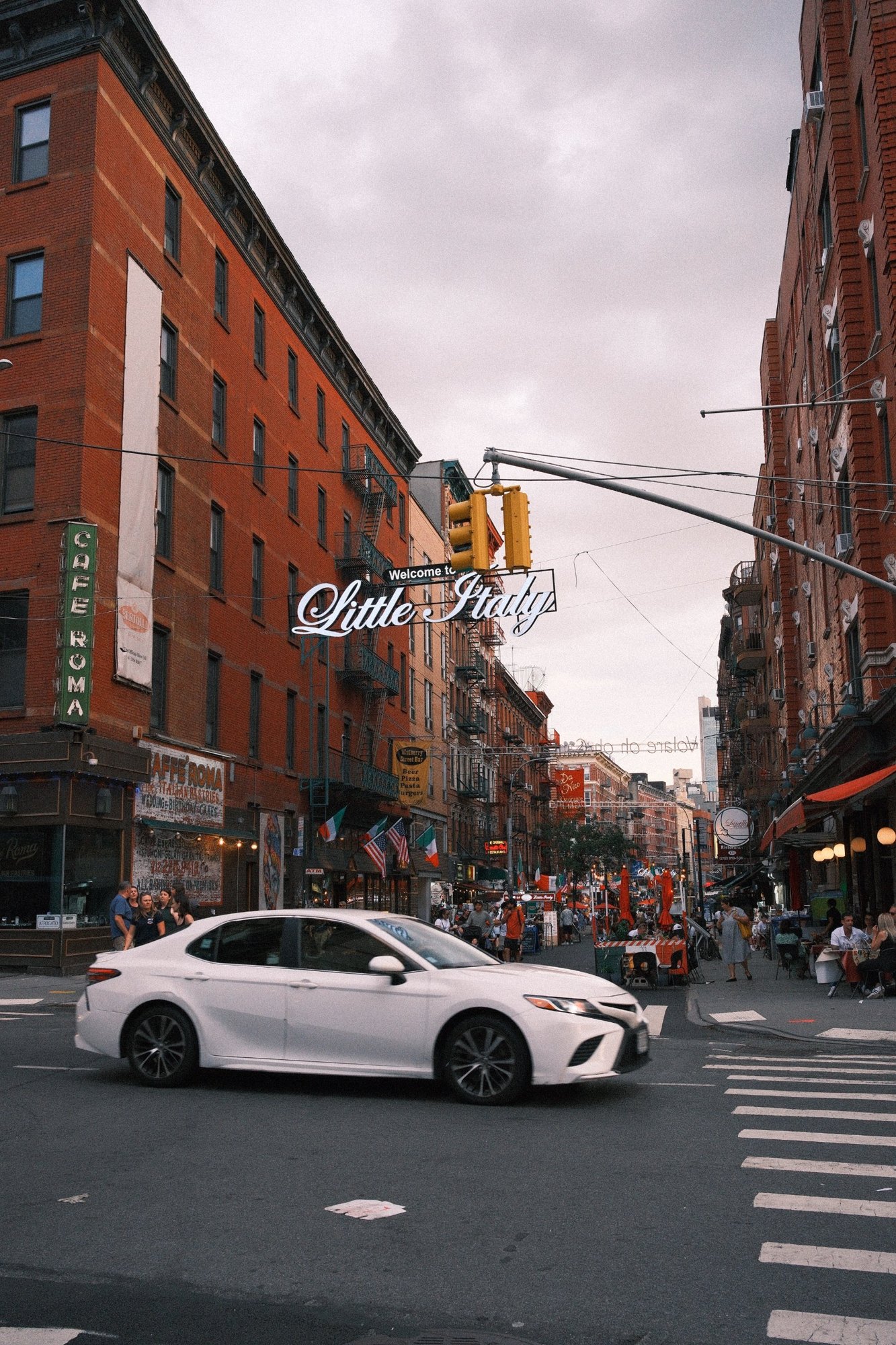 A white car crossing under the Welcome to Little Italy sign
